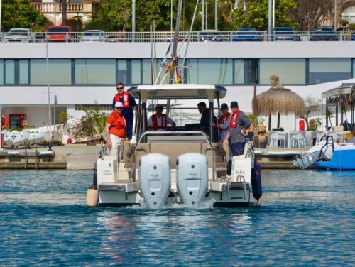 Trainingsgruppe mit roten Schwimmwesten auf einem Motorboot beim Ablegen im Hafen Port Calanova, mediterrane Hafenpromenade im Hintergrund